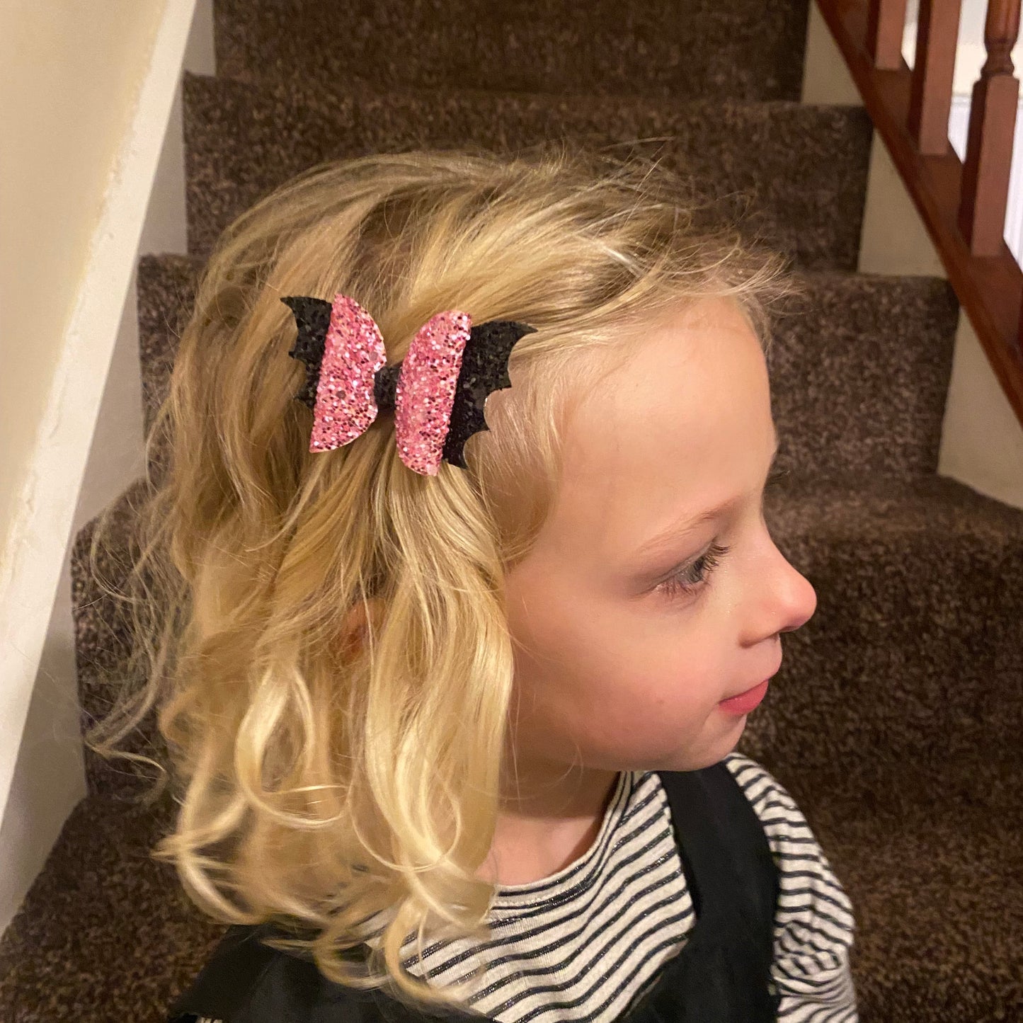 A girl with blonde curly hair wearing a pink glitter vampire hair bow with sparkly black bat wings, sitting on a carpeted staircase.