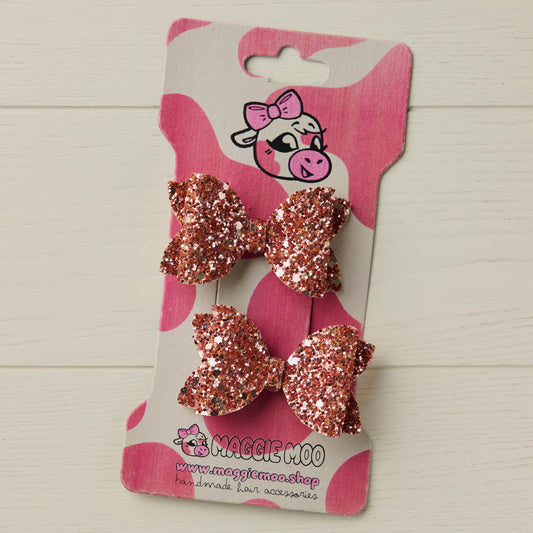 Two rose gold glitter hair bows on a display card with maggie moo logo, on a light wooden surface.