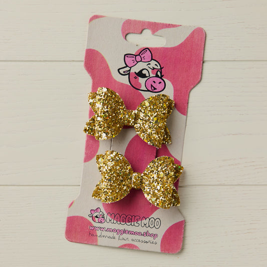 Gold glitter hair bows on a Maggie Moo branded card against a light wooden background