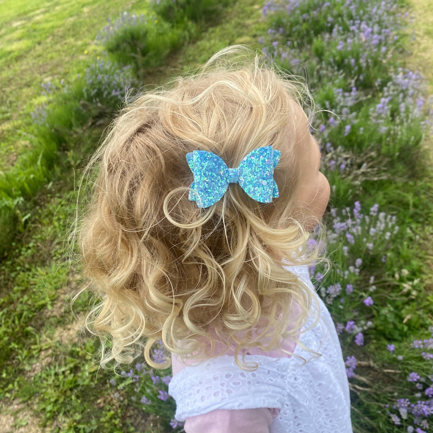 Close-up of a girl with curly blonde hair wearing a sparkly blue hair bow, standing in a field with flowers.
