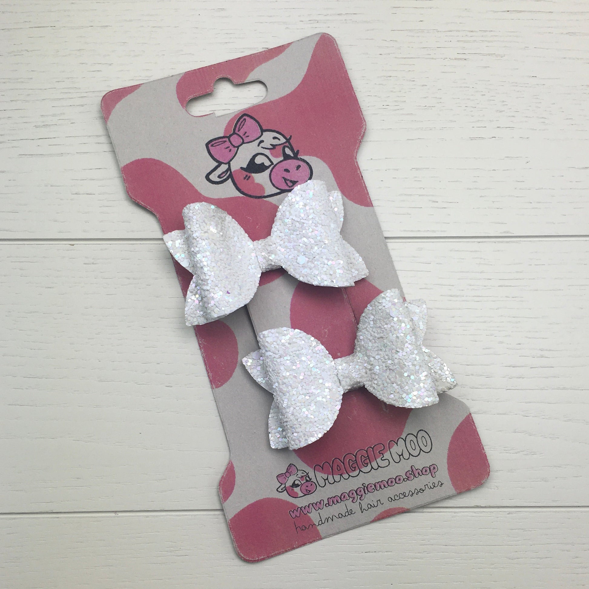 Two white glitter hair bows on a display card with a cow logo on a light wooden surface.