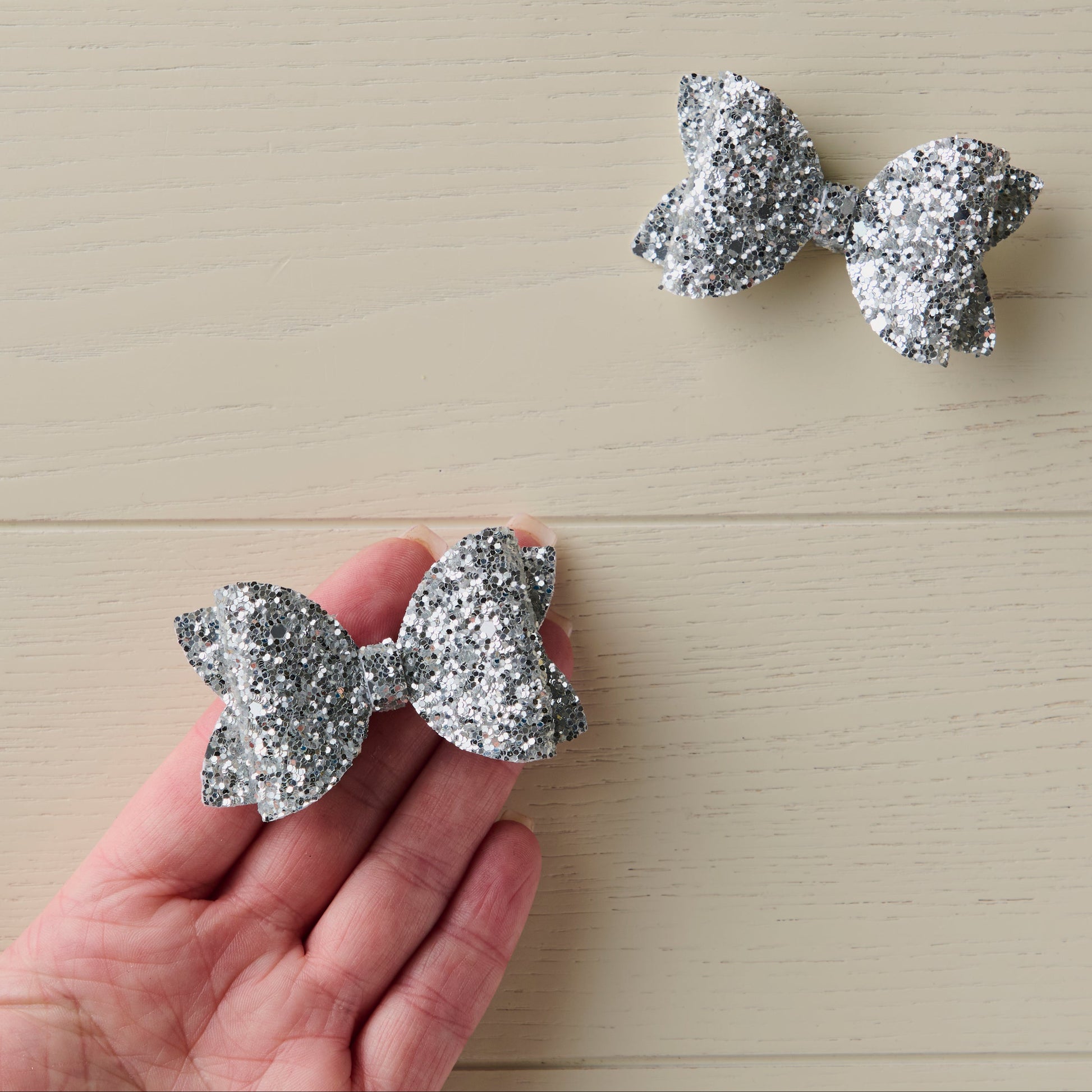 Silver glitter bows on a light wooden surface with a hand holding one for size reference