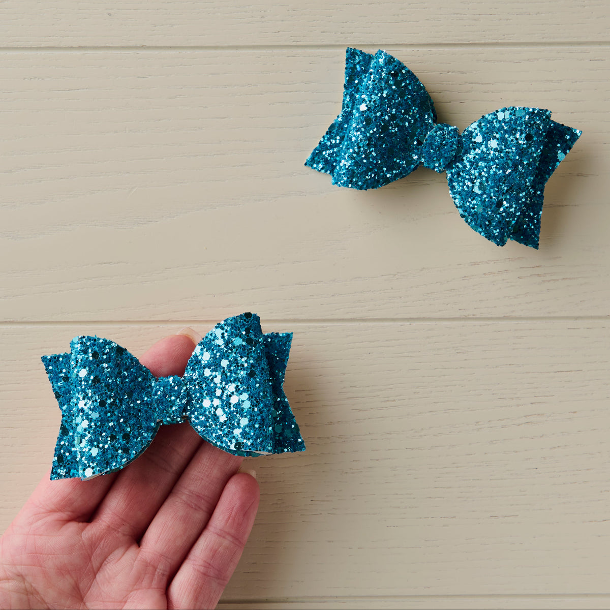 Two blue glittery bows on a light wooden background with one being held on a palm of a hand for size reference.