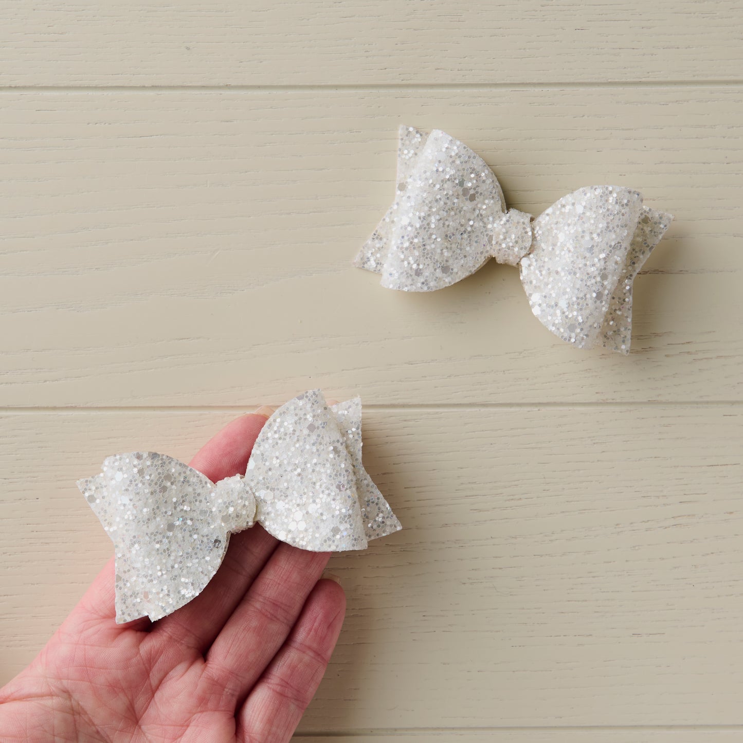 Two glittery white hair bows on a wooden surface with a hand holding one bow.