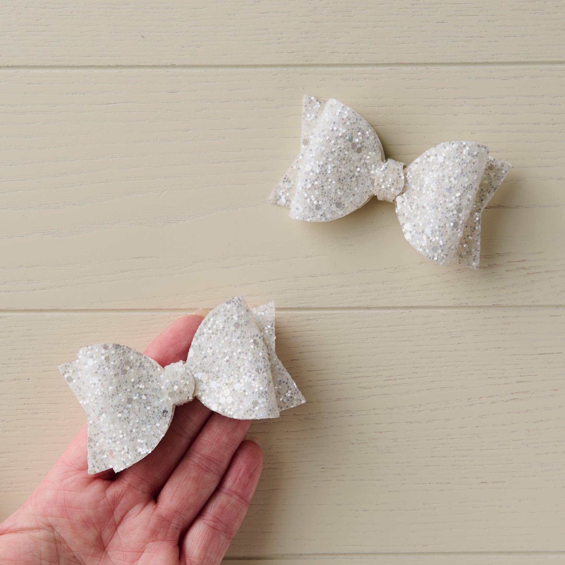 Two glittery white hair bows on a wooden surface with a hand holding one bow.