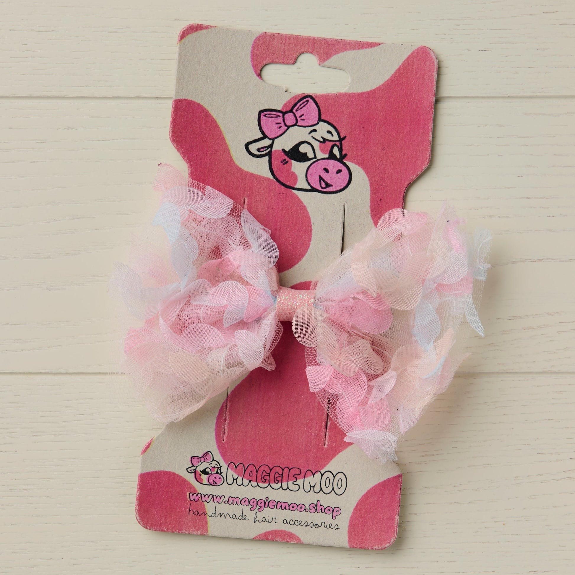 Pink hair bow with 3D floral details on a display card with a cute cow logo against a light wooden background