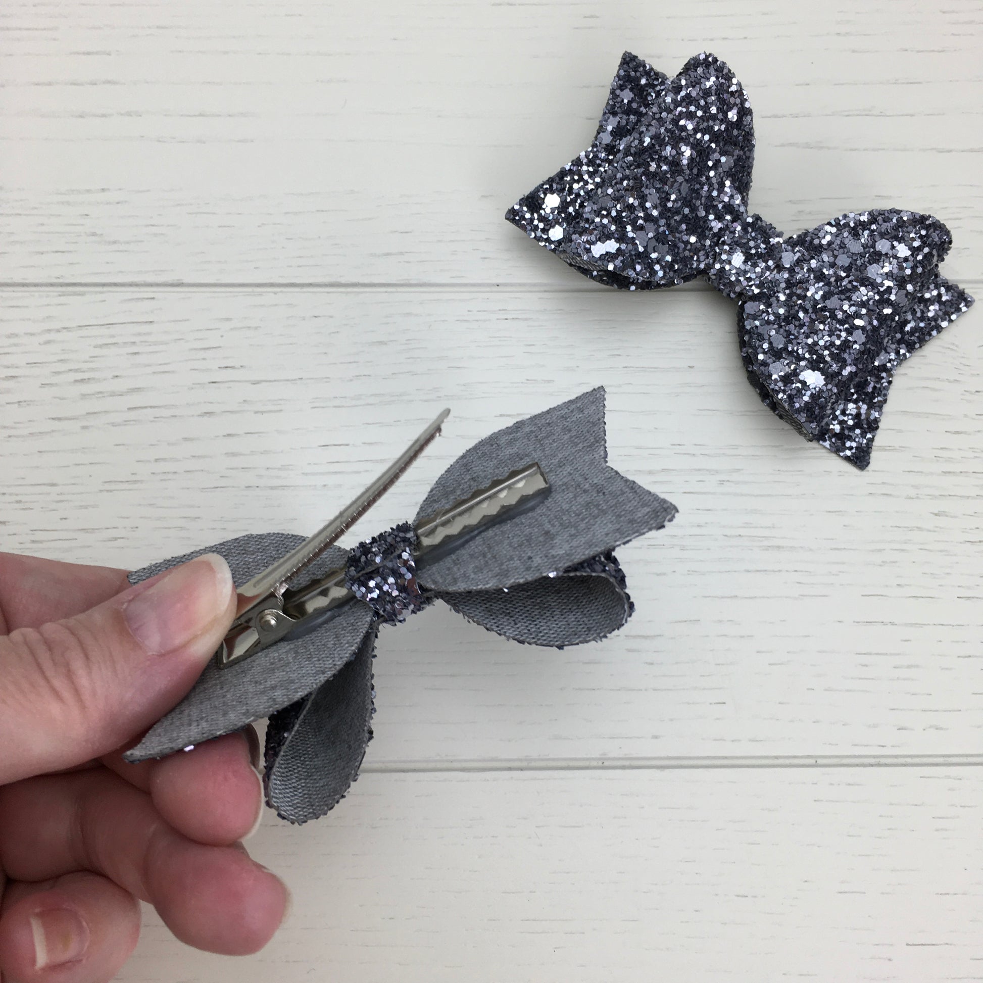 Photo showing the alligator clip on the back of a grey glittery hair bow and the other hair bow on a light wooden surface