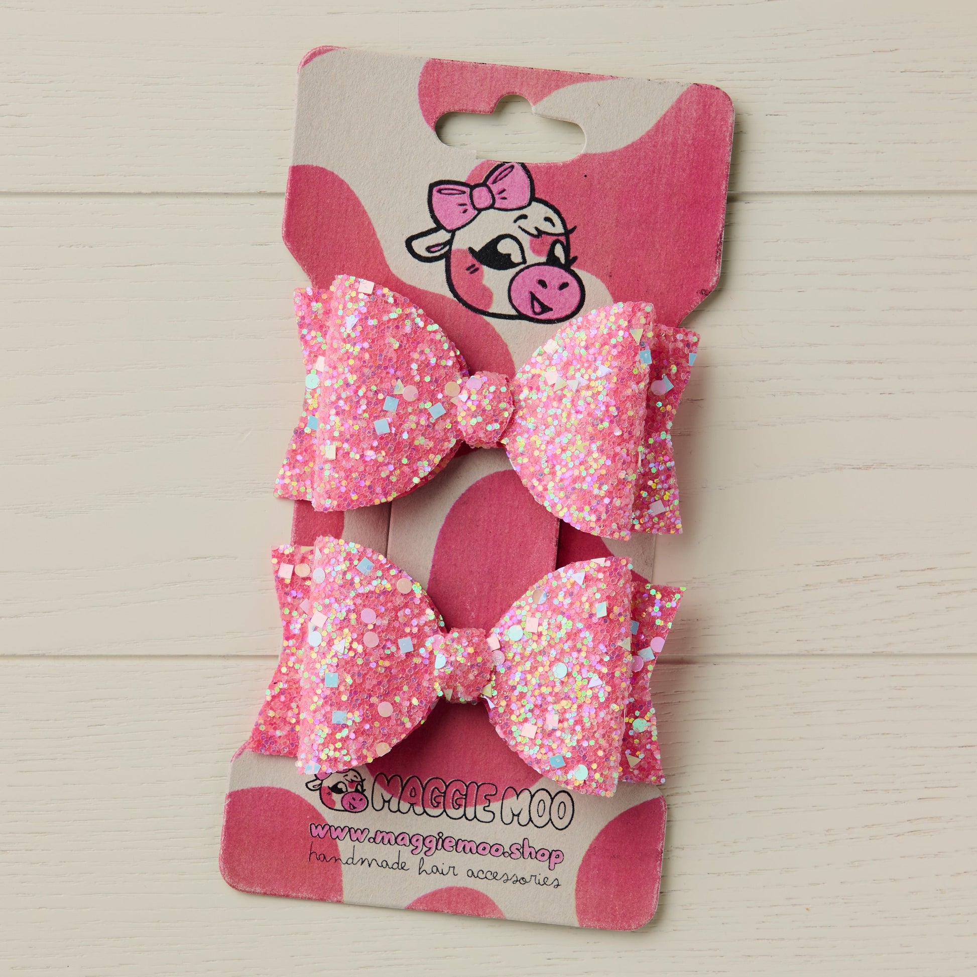 Two pink glittery bows on a display card with a cute cow logo against a light wooden background