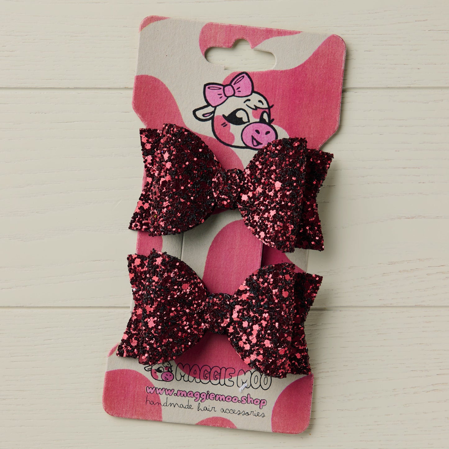 Two dark red glitter hair bows on a display card with a cute cow logo on a light wooden surface.
