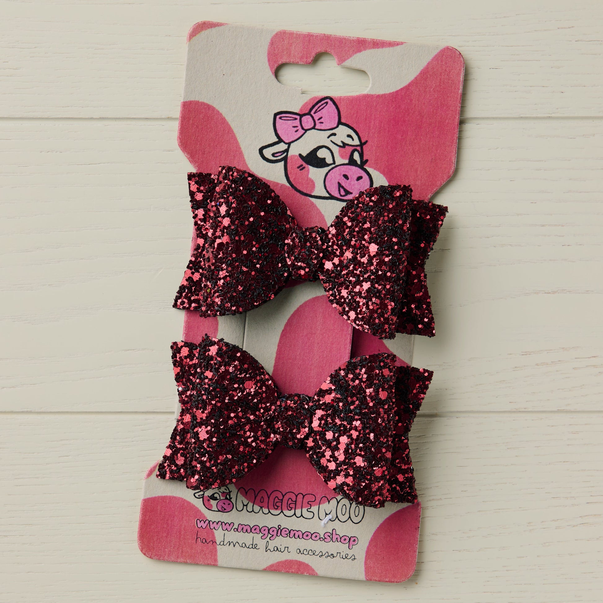 Two dark red glitter hair bows on a display card with a cute cow logo on a light wooden surface.