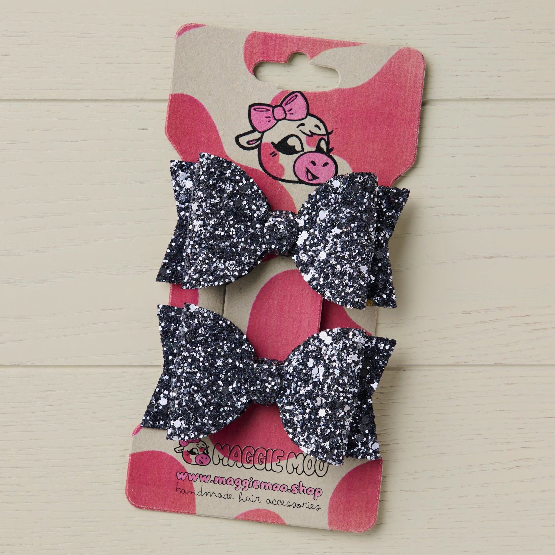 Two glittery grey hair bows on a display card with a cartoon cow logo.