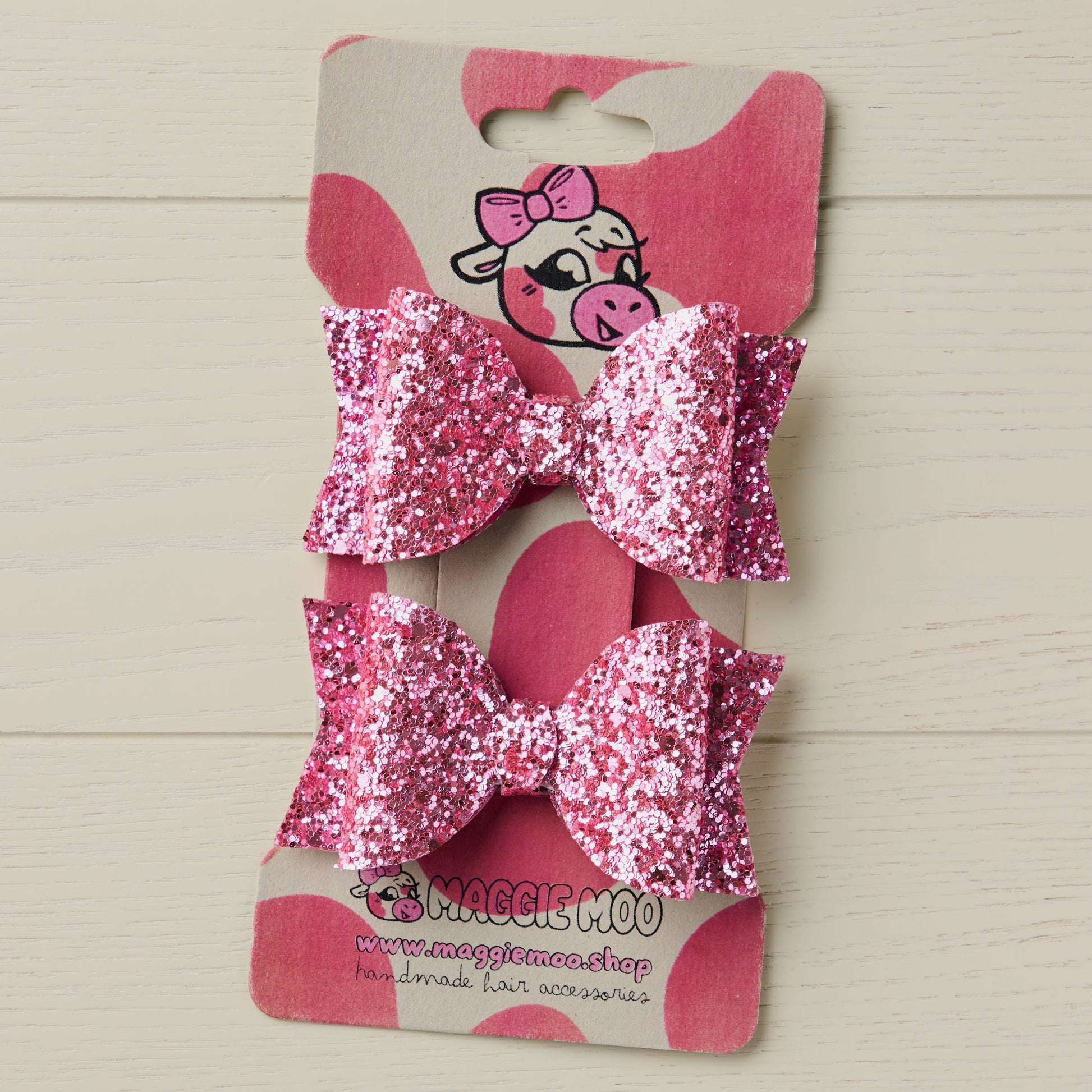 two glitter hair bows on a display card with a cow logo against light wooden background.