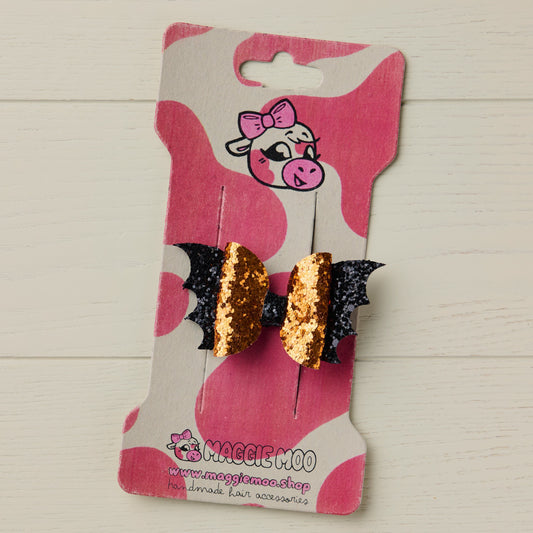 A handmade mini orange and black glitter Halloween hair bow with spooky bat wings, presented on a Maggie Moo display card with a cute cow logo.