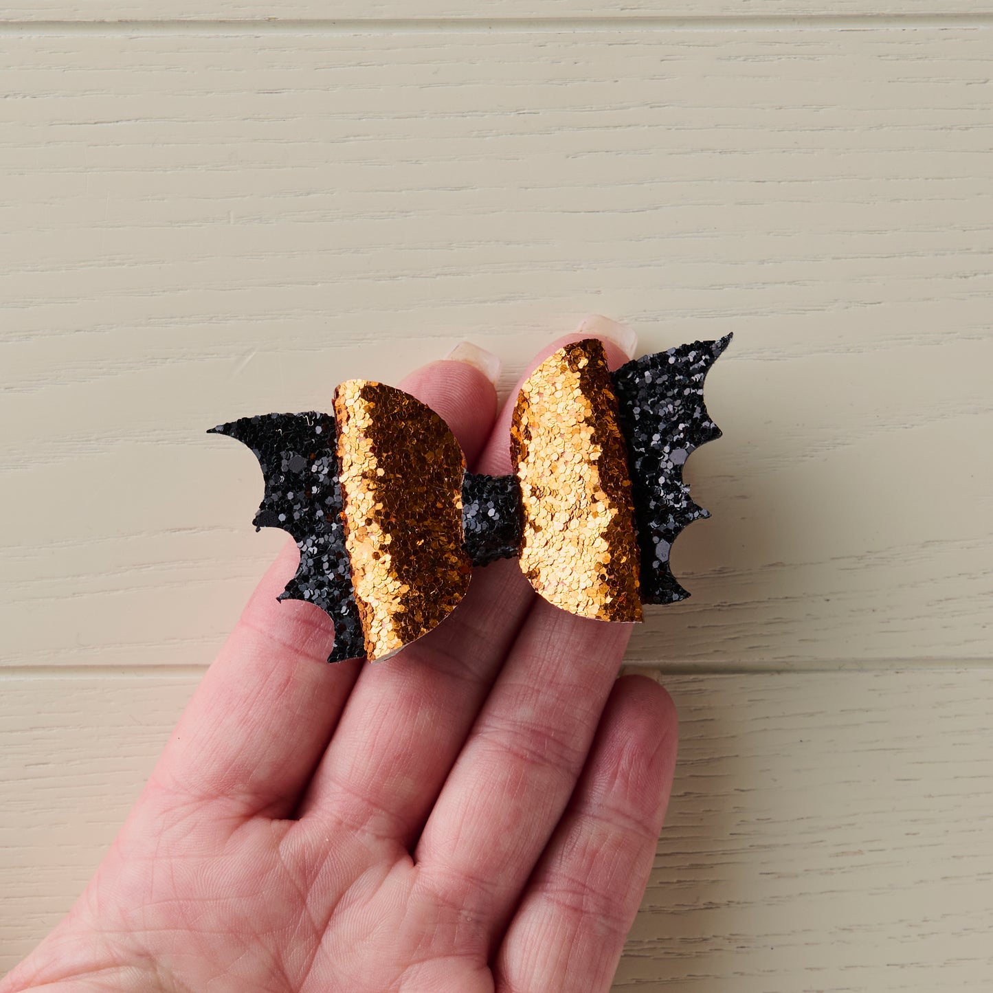 A close-up photo of a small 2.5-inch orange and black sparkly Halloween bat bow being held in a human hand for size reference.