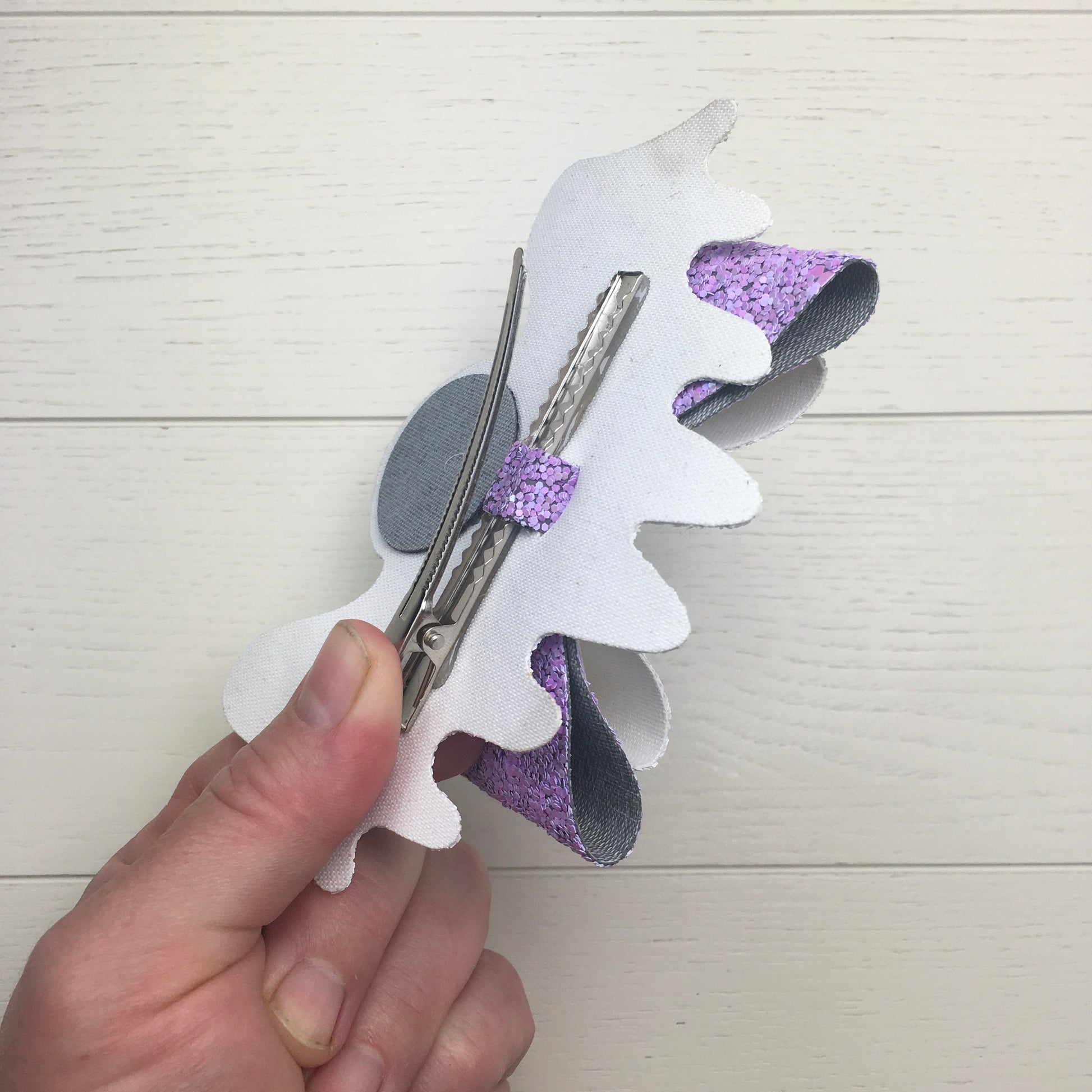 The reverse side of a large glitter Halloween hair bow with a human hand holding the 75mm alligator clip open to show the secure attachment with the centre strip.