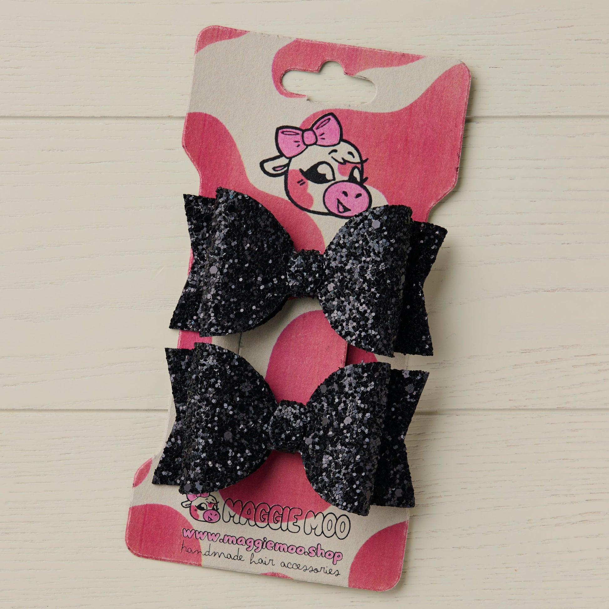 two 3 inch black glitter hair bows on a display with a cute cow logo on a light wooden background