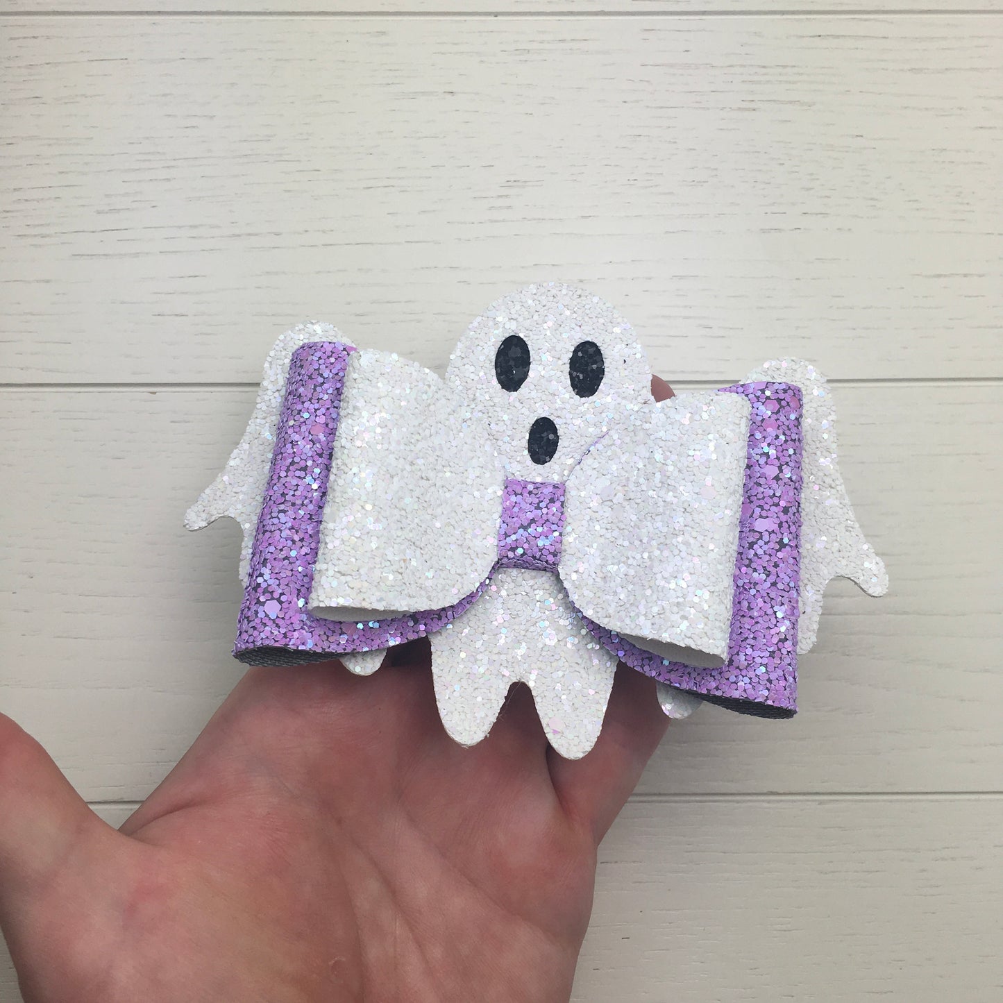 A close-up photo of a large 3 by 4 inch lilac and white sparkly ghost hair bow held in a human hand to show the actual size.