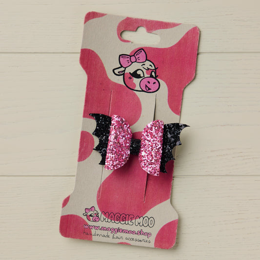 A handmade mini pink and black premium jazz glitter Halloween hair bow with bat wings, on a Maggie Moo display card with a cute cow logo.
