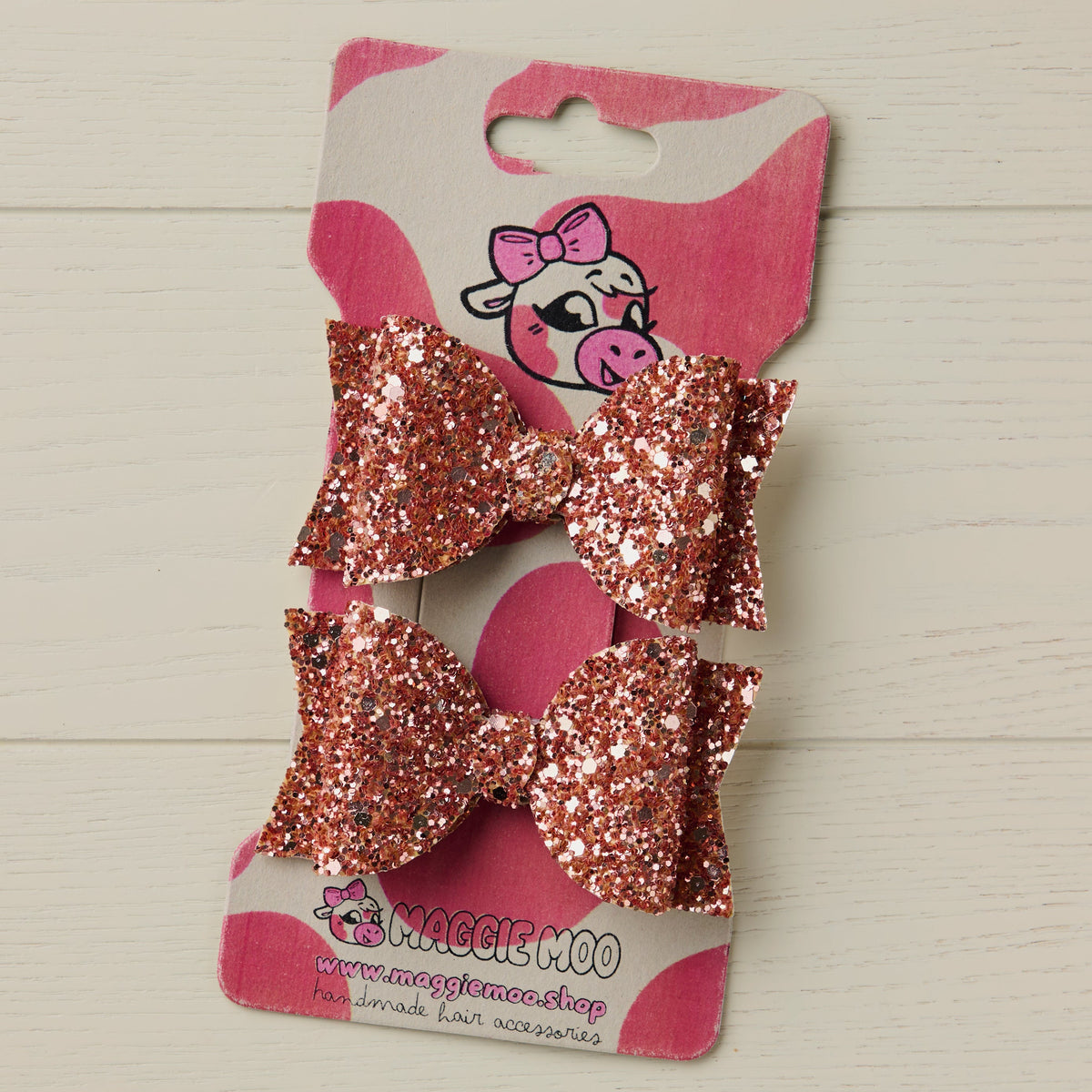 Rose gold glitter hair bows on a display card with a cute cow logo on a light wooden surface