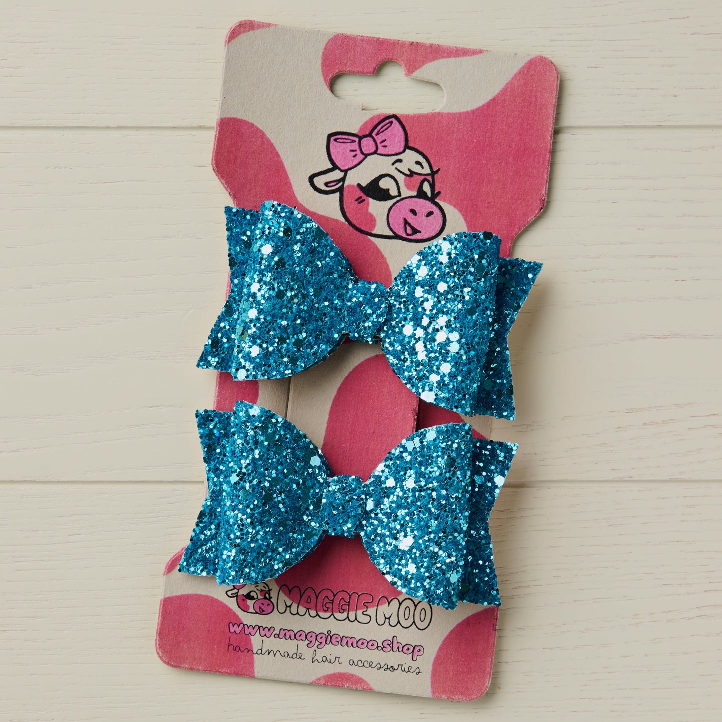 Two sparkly blue jazz glitter hair bows on a display card with a cartoon cow logo on a wooden surface.