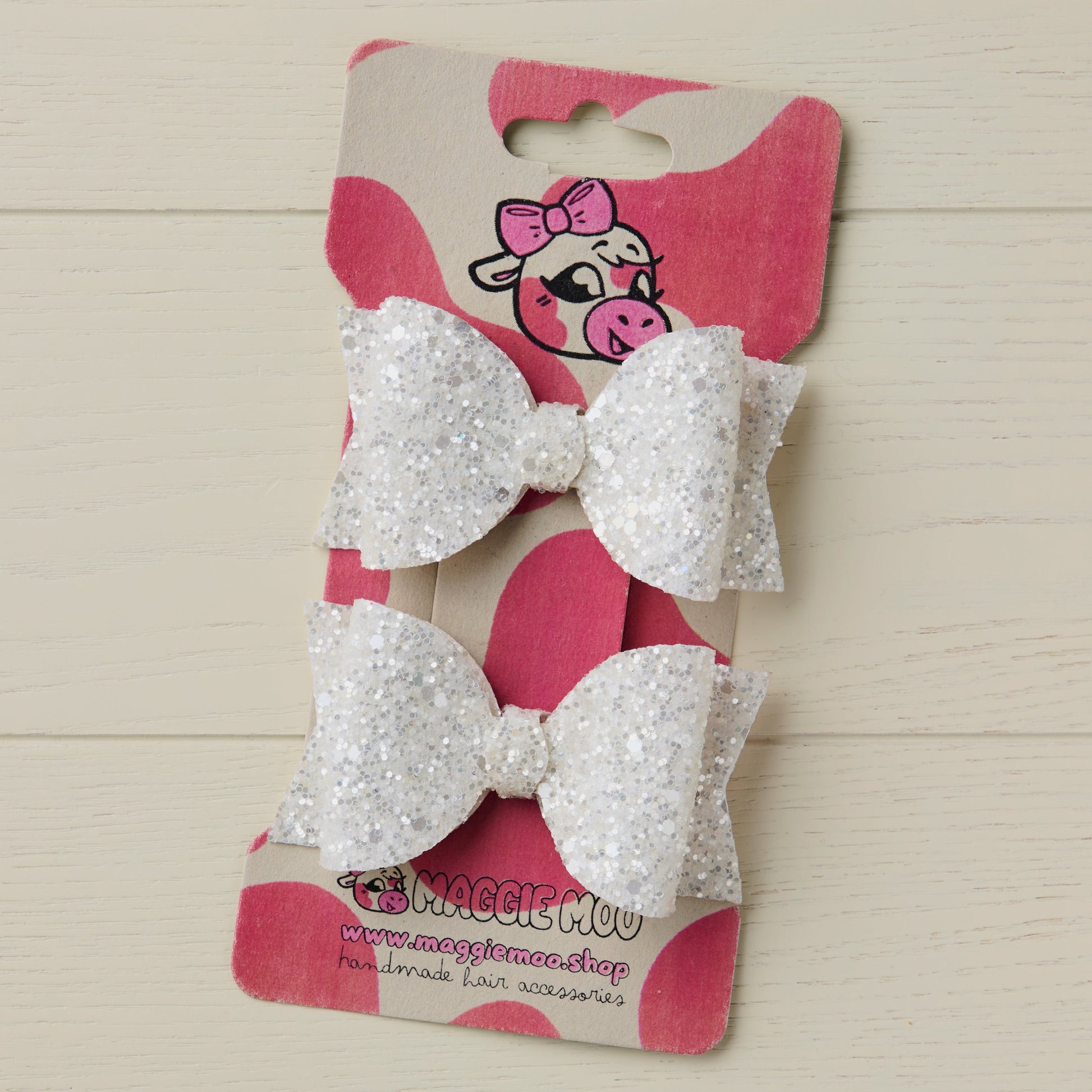 Two pearl white glitter hair bows on a display card with a cute cow logo on a light wooden background.